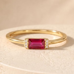 May include: A gold ring featuring a rectangular baguette-cut vibrant pink gemstone set horizontally. The center stone is flanked on both sides by a small cluster of three tiny clear round accent stones embedded into the gold band.