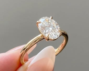 May include: A gold ring featuring a single oval-cut clear gemstone set in a four-prong setting. The band is thin and polished, showcasing a warm metallic tone that complements the brilliant, faceted white stone held securely at the center.