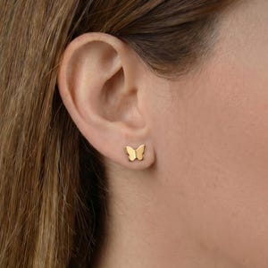 Peut inclure: Une petite boucle d'oreille à clou en forme de papillon de couleur dorée avec une finition lisse et polie. La boucle d'oreille représente un papillon aux ailes symétriques, portée sur le lobe de l'oreille, au design simple et métallique.