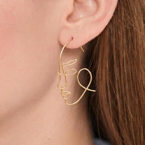 May include: A gold-toned earring crafted from a single continuous wire, shaped into the abstract outline of a human face in profile. The design features a curved line forming the forehead, nose, and chin, with a loop representing the eye area.
