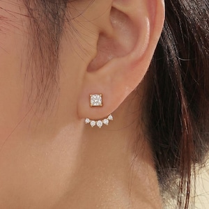 May include: A pair of gold-toned earrings. Each earring features a square stud with a clear stone, and a curved row of smaller, clear stones that sit behind the earlobe. The earrings are displayed on a person's ear.