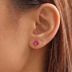 May include: A small stud earring featuring a vibrant, heart-shaped pink gemstone set in a gold-toned metal prong setting. The earring is positioned securely in the earlobe, showcasing a polished finish and a classic, minimalist design aesthetic.