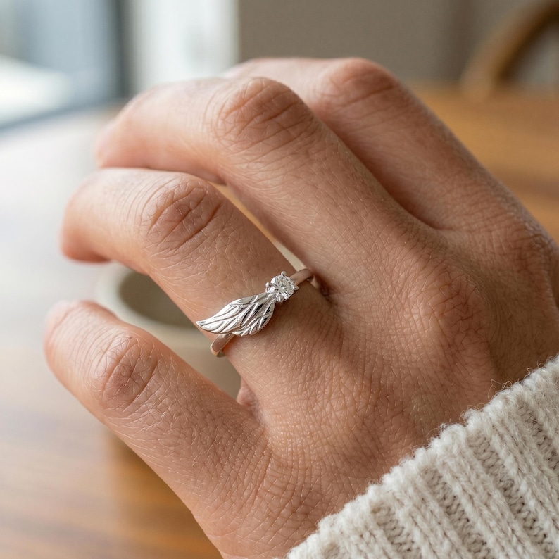 Silver angel wing diamond ring displayed in a soft pink jewelry box with warm bokeh lighting, featuring a detailed wing design beside a round center stone on a slim polished silver band. A beautiful ring for promise, anniversary, bridal jewelry, daily wear, angel inspired jewelry, and fine jewelry gifting. Perfect for buyers looking for a silver wing ring with clean sparkle.