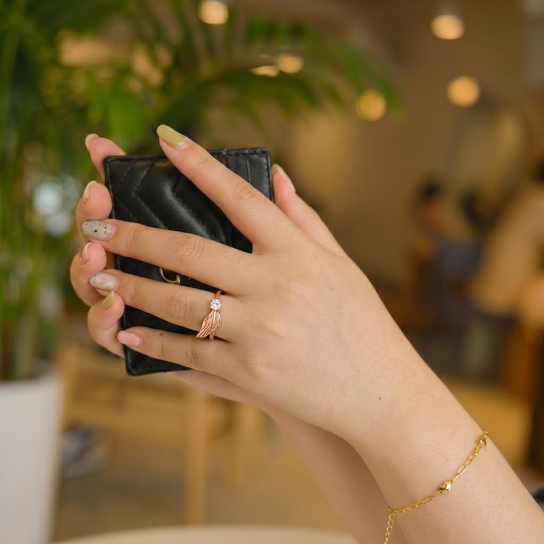 May include: A rose gold ring with a feather design and a clear, round gemstone. The ring is on a hand holding a black, quilted wallet. A gold bracelet with small beads is also visible.