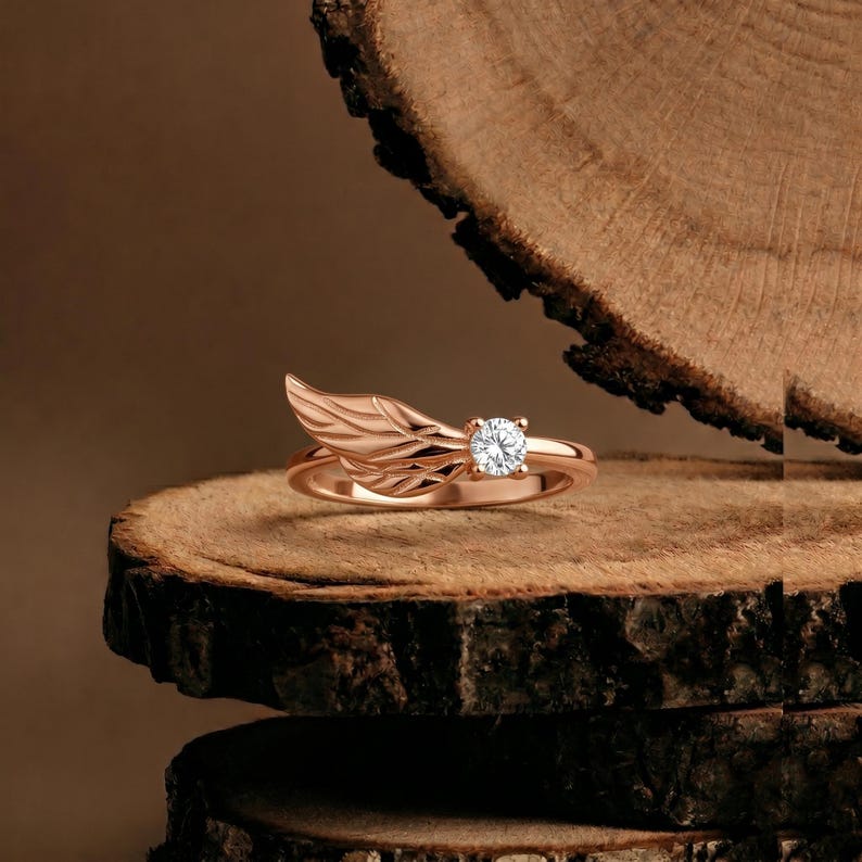May include: Rose gold ring featuring a wing design on one side and a round, clear gemstone on the other. The ring is set on a wooden surface, showcasing its intricate details and elegant style.