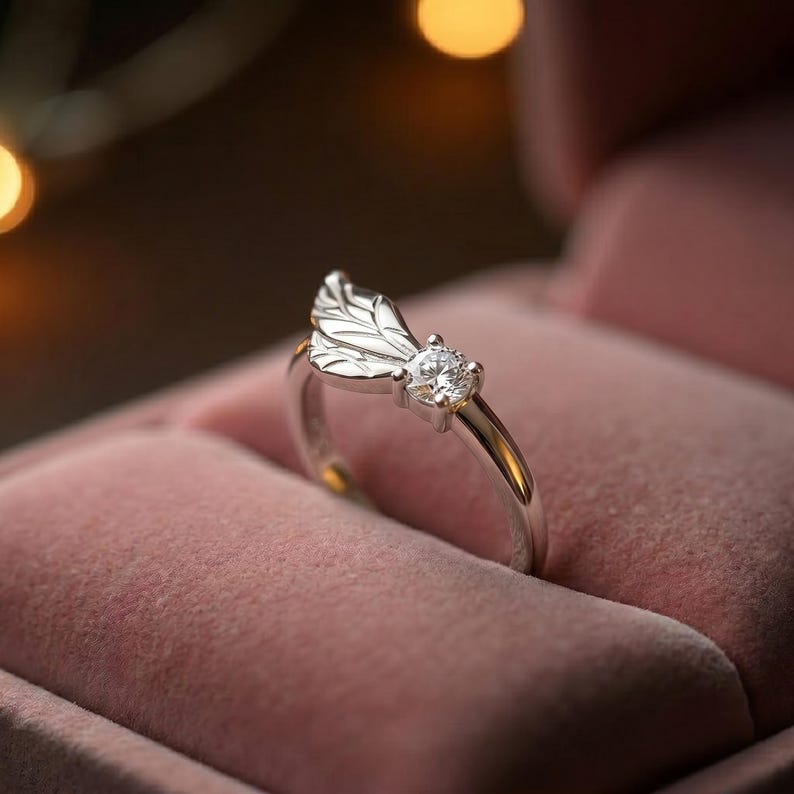 Silver angel wing diamond ring displayed in a soft pink jewelry box with warm bokeh lighting, featuring a detailed wing design beside a round center stone on a slim polished silver band. A beautiful ring for promise, anniversary, bridal jewelry, daily wear, angel inspired jewelry, and fine jewelry gifting. Perfect for buyers looking for a silver wing ring with clean sparkle.