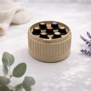 Può includere: Un contenitore beige per oli essenziali che contiene diverse piccole bottiglie scure con etichette colorate. Il contenitore è rotondo con scanalature verticali. Lo sfondo include rametti di lavanda ed eucalipto.
