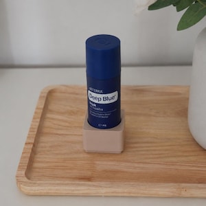 Könnte beinhalten: Ein zylindrischer blauer dōTERRA Deep Blue Stick mit Copaiba-ätherischem Öl-Blend, mit weißem Text auf dem Etikett und einem Nettogewicht von 48g. Das Produkt steht aufrecht in einem kleinen, quadratischen, hellbeigen Halter.