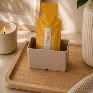 Könnte beinhalten: Ein gelber, rechteckiger Nahrungsergänzungsmittelbeutel mit der weißen Aufschrift dōTERRA xEO Mega+, der aufrecht in einem kleinen, hellbeigen, rechteckigen Kunststoffbehälter steht. Der Beutel ist mit einem vertikalen weißen Streifen versehen.
