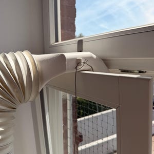 Könnte beinhalten: Ein weißer Kunststoff-Fensteradapter für Klimageräte mit einem flexiblen Akkordeon-Schlauch, der an einer starren Düse befestigt ist. Die Düse ist mit einer dünnen Metalldrahtschlaufe an einem kleinen Kunststoffhaken am weißen Fensterrahmen gesichert.