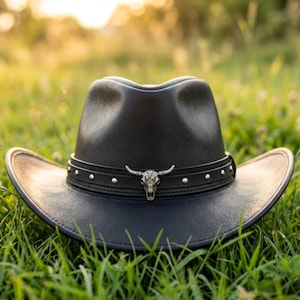May include: A black leather cowboy hat with a decorative band featuring silver studs and a silver bull skull emblem. The hat is resting on green grass, with a blurred background.