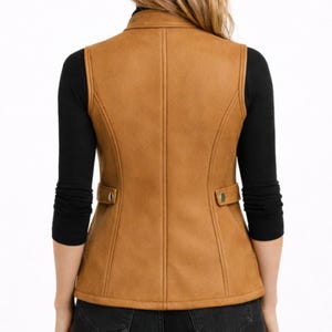 May include: A tan vest with a collared neckline and two front pockets. The vest has a tailored fit and a button closure on each pocket. The back of the vest is visible, showing vertical seams and a structured design. The vest is worn over a black long-sleeved shirt.