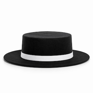 May include: A black hat with a flat top and brim, featuring a white band around the crown. The hat has a classic design, suitable for various occasions. The hat's brim is wide, and the hat is set against a white background.