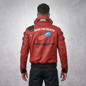 Könnte beinhalten: Rote Lederjacke mit schwarzen Akzenten, mit dem Text "AKIRA" am Kragen und "GOOD FOR HEALTH BAD FOR EDUCATION" mit einer Pillengrafik auf dem Rücken. Die Jacke hat einen Bomberstil mit schwarzem Rippsaum und Bündchen.