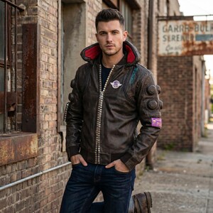 Könnte beinhalten: Braune Lederjacke mit rot gefütterter Kapuze, silbernem Reißverschluss und mehreren kreisförmigen Akzenten. Die Jacke hat Aufnäher mit Text und einem Logo. Die Person trägt eine blaue Jeans.