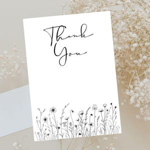 May include: A white card with the words "Thank You" in elegant black script. The bottom of the card features a delicate black and white floral illustration. A blank white envelope is visible to the left.