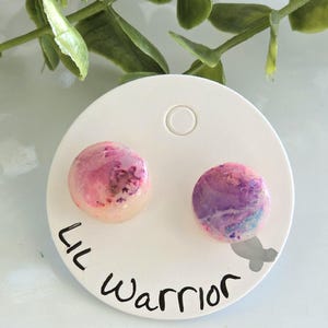 May include: A pair of round stud earrings with a marbled design in shades of pink, purple, and white. The earrings are displayed on a white card with the text "Lil Warrior" in black script.