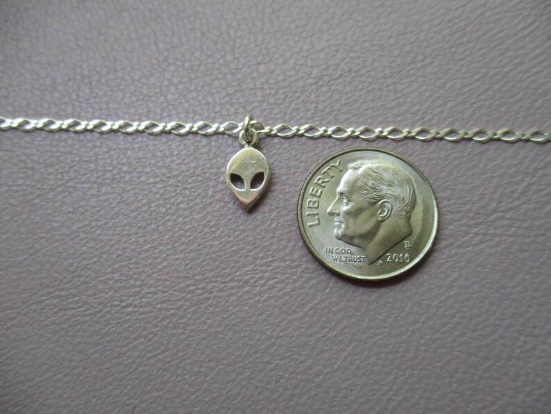 Puede incluir: Un collar de cadena de plata con un peque&ntilde;o dije de extraterrestre. El dije es del tama&ntilde;o de una moneda de diez centavos.