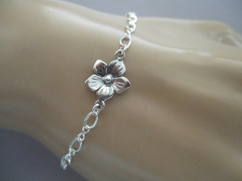 May include: A silver chain bracelet with a delicate flower charm. The flower charm has a detailed design and is attached to the chain with a small loop.