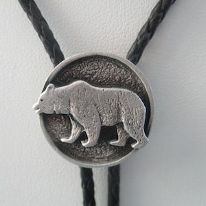 May include: Silver-toned bolo tie featuring a detailed bear design. The bear is centered on a textured, circular pendant, with a braided black cord. A classic accessory for a rustic or Western-inspired look.