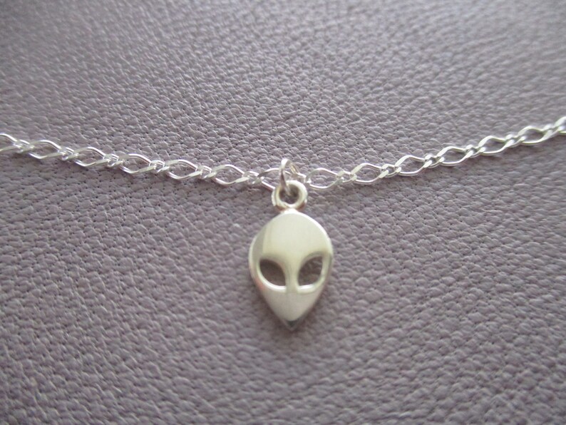 Puede incluir: Un collar de cadena de plata con un peque&ntilde;o colgante de cabeza de extraterrestre de plata.