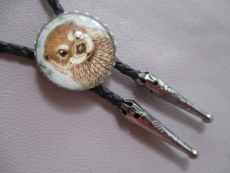 May include: A black bolo tie with a silver-toned metal tip and a round, glass pendant featuring a close-up image of an otter.