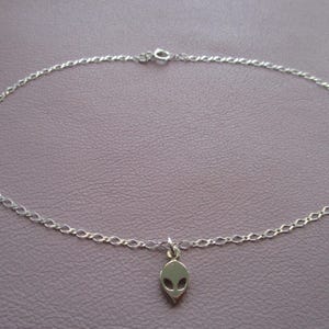 Puede incluir: Un tobillo de cadena de plata con un dije de cabeza extraterrestre.