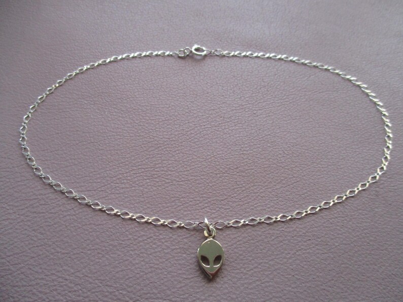 Puede incluir: Un tobillo de cadena de plata con un dije de cabeza extraterrestre.