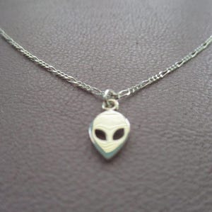 May include: A silver-toned necklace featuring a pendant shaped like an alien head. The pendant has a white face with black eyes and a light blue outline. The chain is a delicate, linked design.