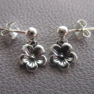 May include: A pair of silver dangle earrings with a small flower charm. The earrings have a post back closure.