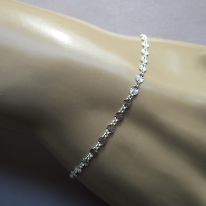 May include: Delicate silver bracelet featuring a repeating floral design. The bracelet is shown on a light-toned arm, showcasing its dainty and elegant appearance. A simple and stylish piece of jewelry.