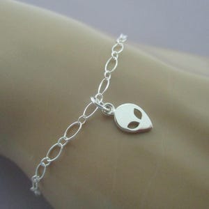 May include: A silver bracelet with an oval link chain and a small alien head charm. The charm has two eye cutouts and a smooth, polished finish. The bracelet is shown on a light skin tone.