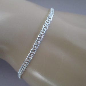 May include: A silver chain bracelet with a delicate, interwoven design. The bracelet is shown on a person's wrist.