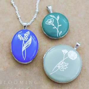 May include: Three silver-toned metal lockets with white floral line art. One oval locket is bright blue on a silver chain. Two round lockets are teal and sage green. The text "BLOOMING thread" is visible in the bottom left corner of the image.