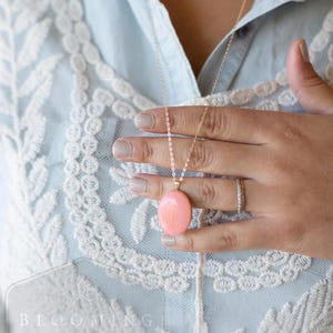 May include: A gold-colored necklace with a large, oval, peach-colored pendant. The necklace is worn over a light blue shirt with white lace detailing. The image also shows a hand wearing a ring.