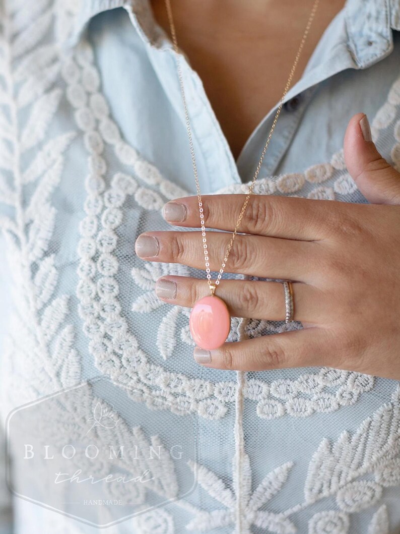 May include: A gold-colored necklace with a large, oval, peach-colored pendant. The necklace is worn over a light blue shirt with white lace detailing. The image also shows a hand wearing a ring.