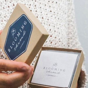 May include: A square cardboard gift box with a lid held open. The lid features a dark blue hexagonal label with the white text "BLOOMING thread" and a flower icon. Inside, a white square card displays the same logo and the word "HANDMADE".