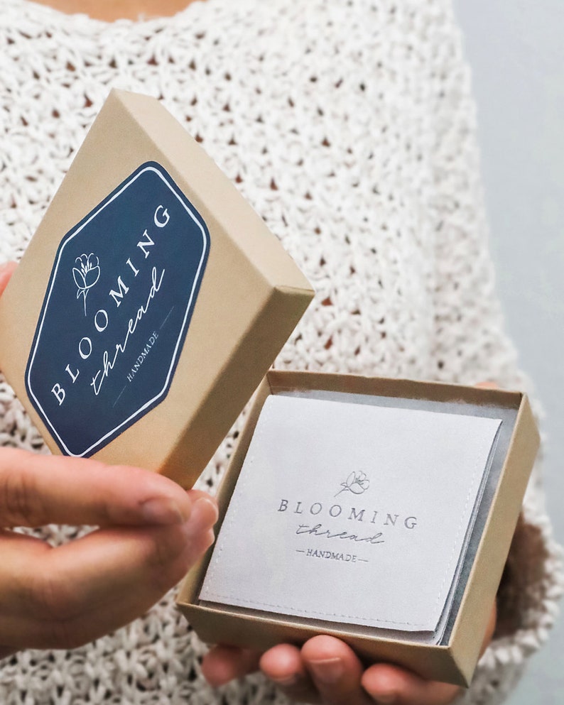 May include: A square cardboard gift box with a lid held open. The lid features a dark blue hexagonal label with the white text "BLOOMING thread" and a flower icon. Inside, a white square card displays the same logo and the word "HANDMADE".