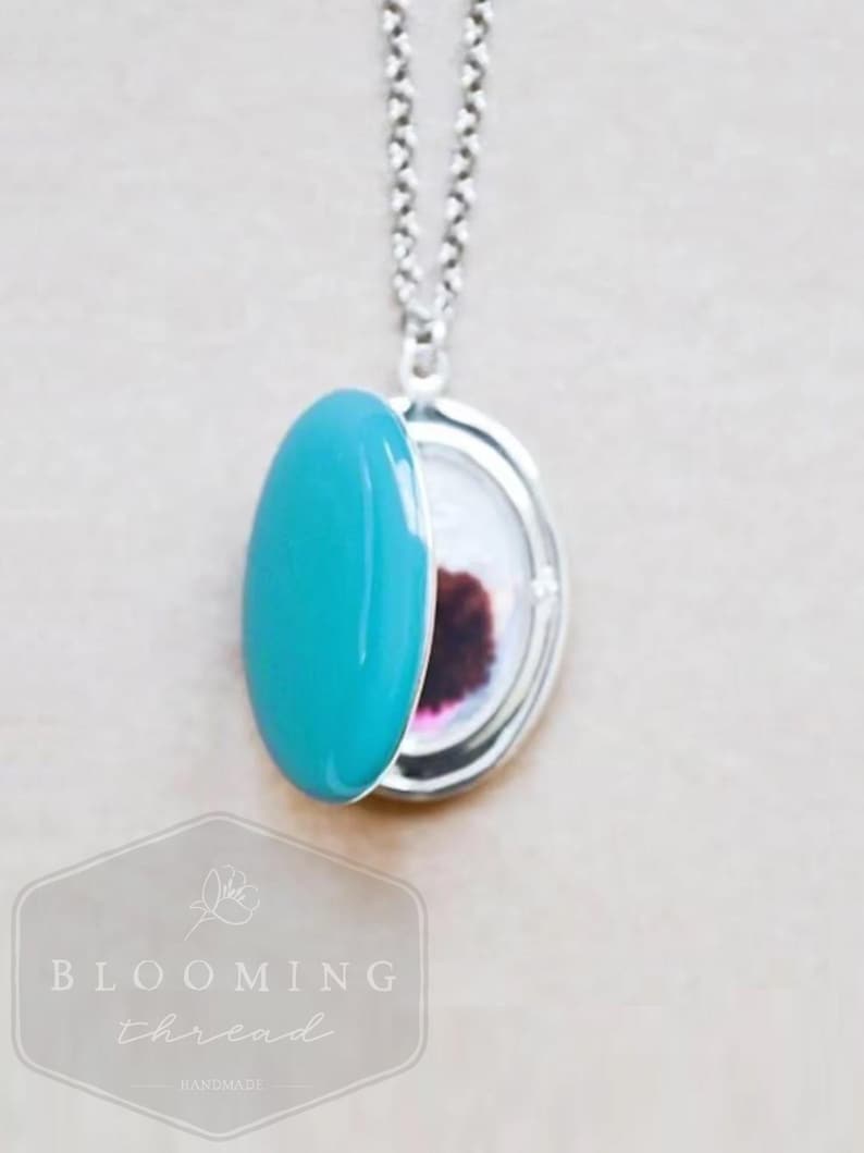 May include: A silver locket necklace featuring a smooth, oval-shaped turquoise enamel cover that is partially open. The interior of the locket reveals a small, circular photograph with a dark, reddish-brown subject. The locket hangs from a silver chain.