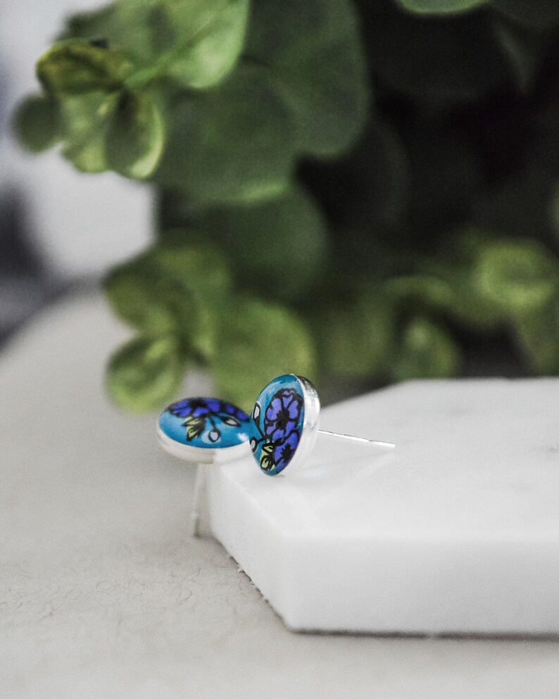 Peut inclure: Une paire de boucles d'oreilles clous en argent avec des motifs floraux bleus et violets sur le devant.