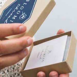 以下が含まれることがあります： 取り外し可能な蓋が付いた正方形の段ボール製ギフトボックス。ダークブルーのラベルには、白い文字で「Blooming Thread」と花のアイコンが印刷されています。箱の中には、同じロゴとテキストが印刷された白い布製のポーチが入っています。