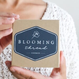 May include: A square brown cardboard box featuring a dark blue hexagonal label with a white floral icon and the text BLOOMING thread HANDMADE printed in white. The box is held in place, showcasing the branding clearly on the front surface.