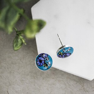 Peut inclure: Une paire de boucles d'oreilles clous argent&eacute;es avec un fond bleu et un motif floral violet.