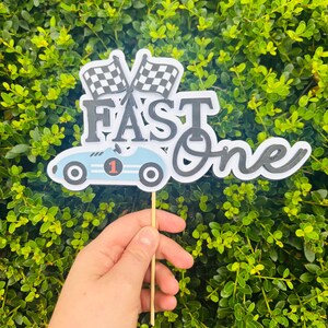 Könnte beinhalten: Hellblau-weiße Tortenaufsatz "Fast One" mit Rennwagen und Zielflaggen. Die Zahl "1" befindet sich auf dem Auto. Der Aufsatz wird von einem Holzstab vor einem grünen, belaubten Hintergrund gehalten.