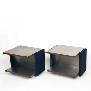 Può includere: Due tavolini moderni dal design minimalista. Ogni tavolo presenta un piano e un ripiano inferiore in metallo spazzolato, con un pannello laterale rivestito in tessuto blu scuro. I tavoli sono su sfondo bianco.