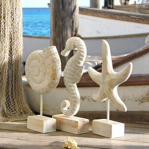 Puede incluir: Tres esculturas costeras de madera: una concha de nautilus, un caballito de mar y una estrella de mar. Cada escultura está montada sobre una pequeña base de madera con una varilla metálica. Las esculturas son de color claro y tienen un aspecto envejecido, evocando un tema de playa.