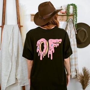 Puede incluir: Camiseta negra con un diseño de donut rosa que dice "OF". El donut tiene detalles amarillos y azules. La persona de la imagen lleva un sombrero marrón.