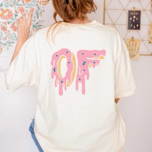 Puede incluir: Camiseta color crema con un gráfico rosa y amarillo de las letras "OF" diseñadas para parecerse a un donut goteando. Las letras tienen chispas azules. La camiseta es de estilo casual y oversize.
