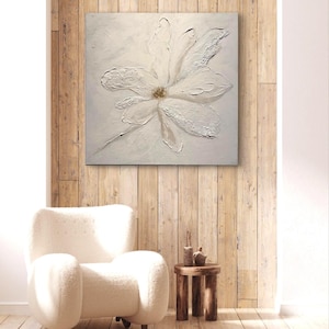 Könnte beinhalten: Ein großes, quadratisches, weißes Blumenbild mit strukturierten Blütenblättern und einem goldenen Zentrum wird an einer Holzwand ausgestellt. Das Kunstwerk ist der Mittelpunkt eines Raumes mit einem weißen Sessel und einem kleinen Beistelltisch aus Holz.