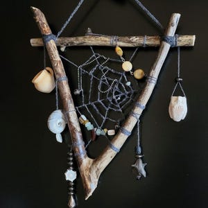 May include: A handmade dreamcatcher with a triangular wooden frame. The frame is made of natural branches and adorned with a gray web, beads, shells, and small carved objects. The dreamcatcher hangs against a black background.